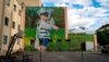 Ein Mural mit einem Jungen mit kurzer blauen Hose und blau-weiß gestreiften T-Shirt vor einem grünen Hintergrund aus Gras und Bäumen. Der Junge hat den Kopf und den Oberkörper in eine Luftpolsterfolie gewickelt.