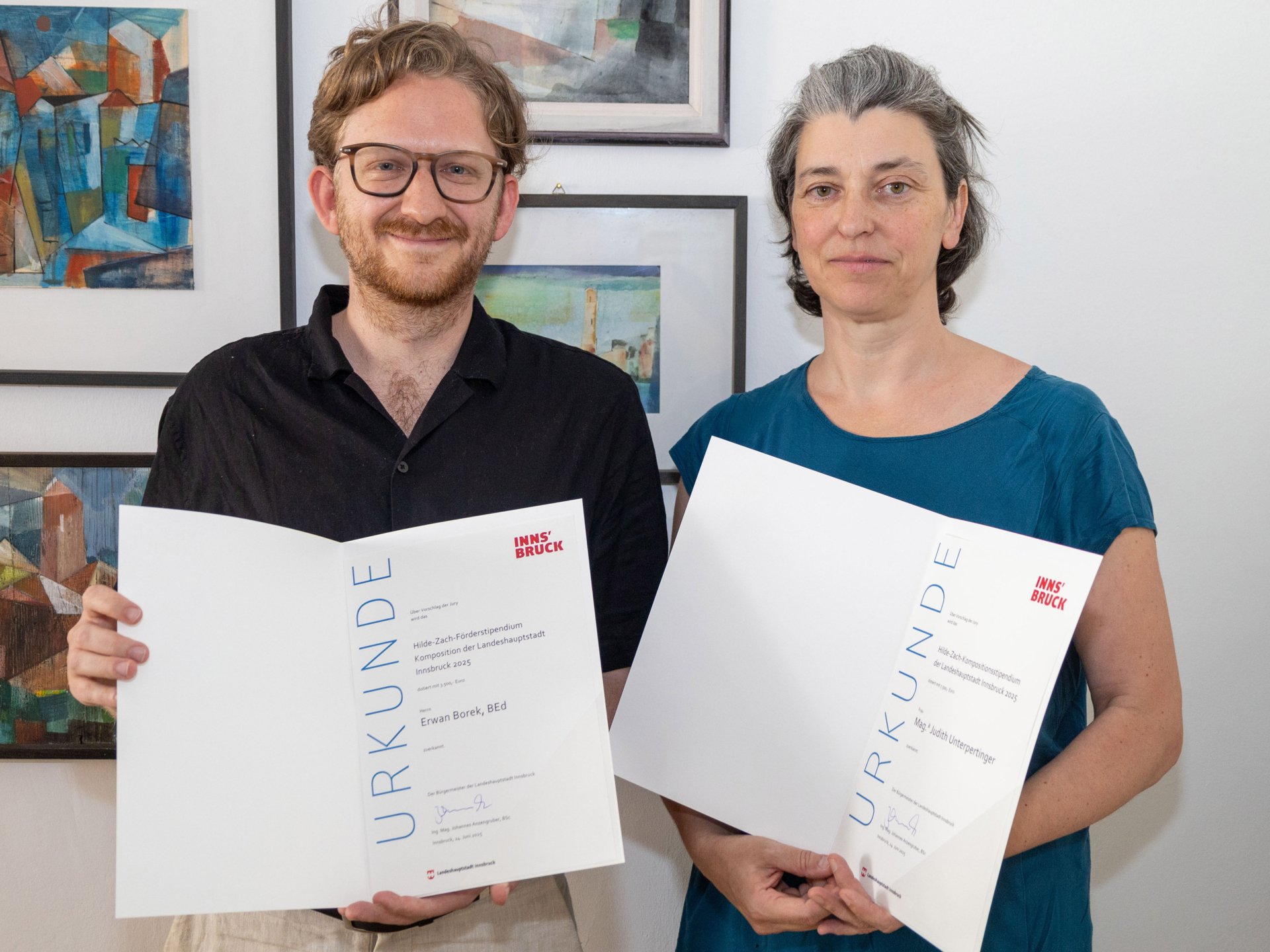 Die Kompositions-StipendiatInnen Erwan Borek und Judith Unterpertinger halten ihre Urkunden in der Hand und lächeln in die Kamera. Dahinter sieht man eine Bilderwand.