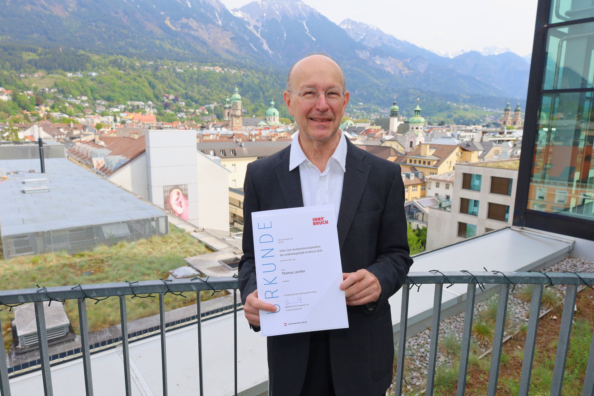 Die Kompositions-StipendiatInnen Erwan Borek und Judith Unterpertinger halten ihre Urkunden in der Hand und lächeln in die Kamera. Dahinter sieht man eine Bilderwand.
